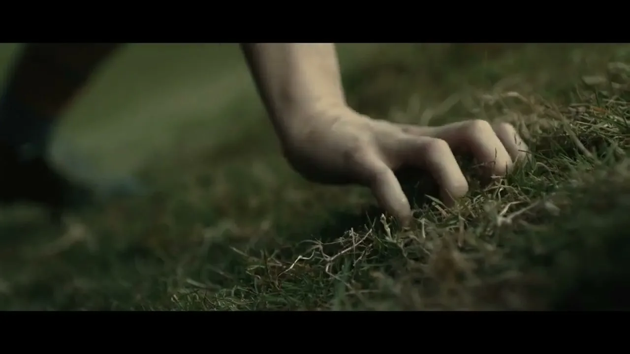 A child's hand with dirt under fingernails grips grass on a field, as a blurry foot steps in the background.