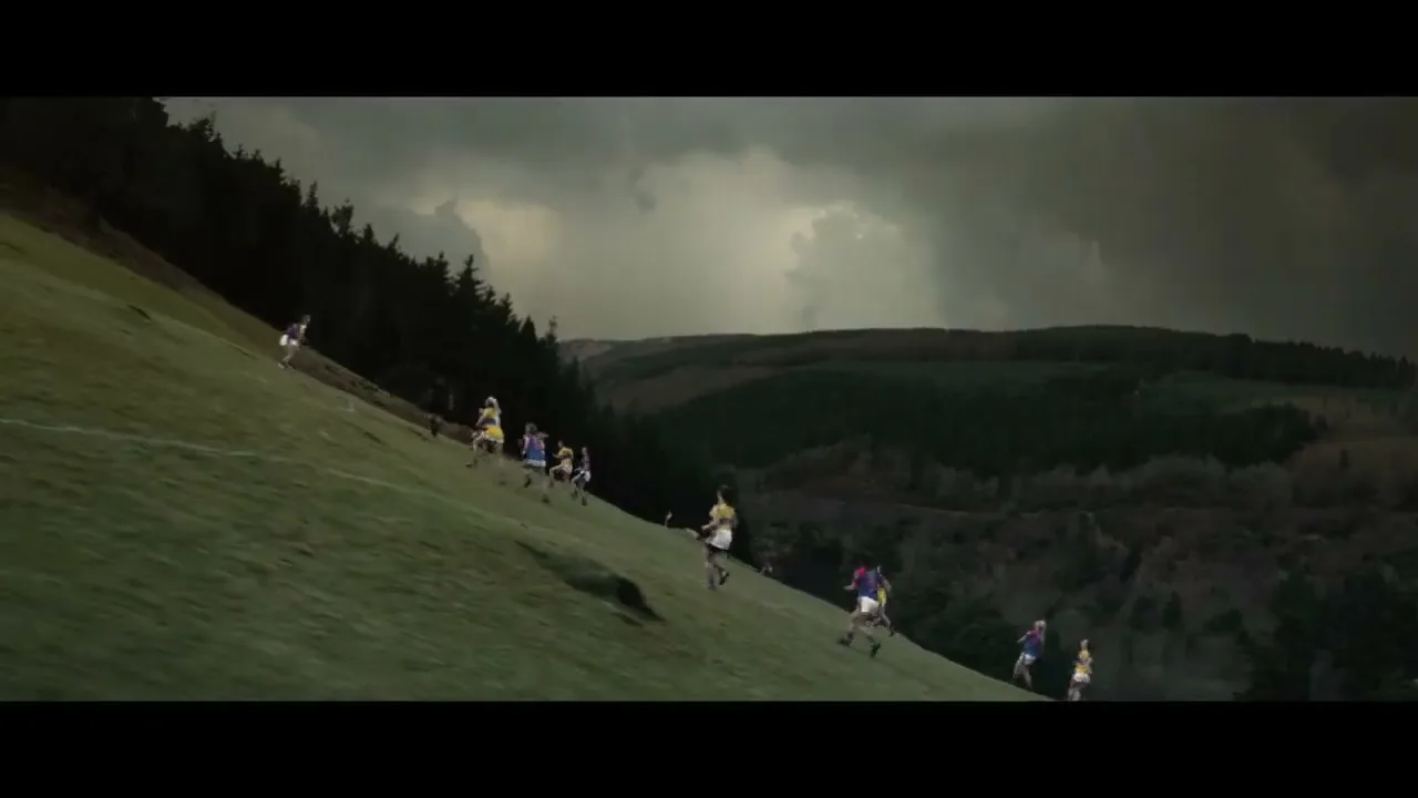 A group of female athletes in colorful jerseys run up a steep grassy hill under a dramatic, cloudy sky, with a forest in the background.