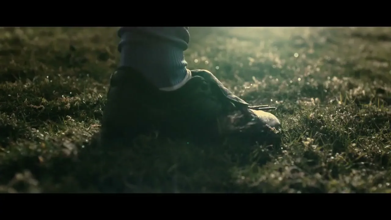 A child's foot in a muddy football boot kicks up mud and grass on a wet, dewy football pitch during the day.