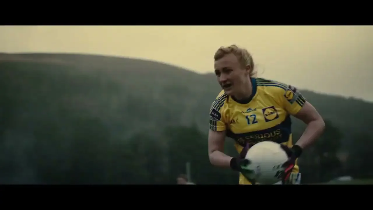 A female Gaelic football player in a yellow and blue jersey runs with the ball across a field with hills in the background under an overcast sky.