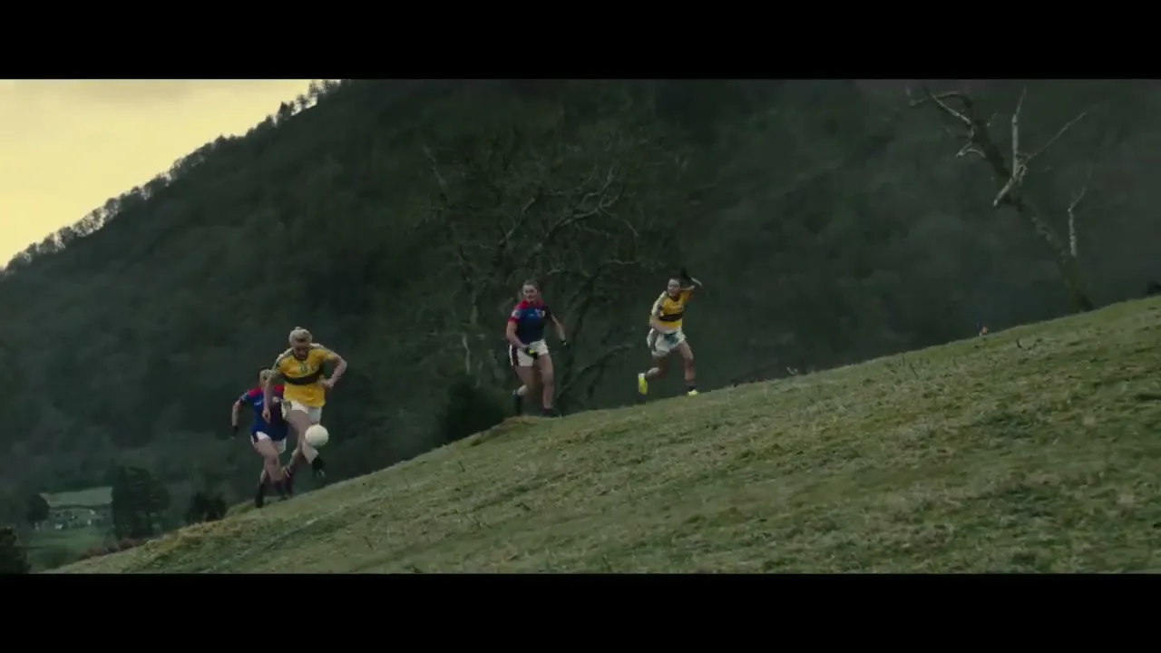Four female Gaelic football players run up a grassy hill with a dark, forested mountain in the background.