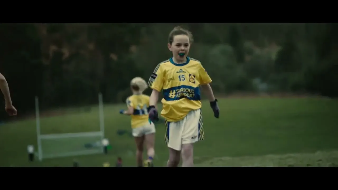 A young girl in a yellow and blue sports jersey with a mouthguard runs on a grassy field during a game, with other players in the background.