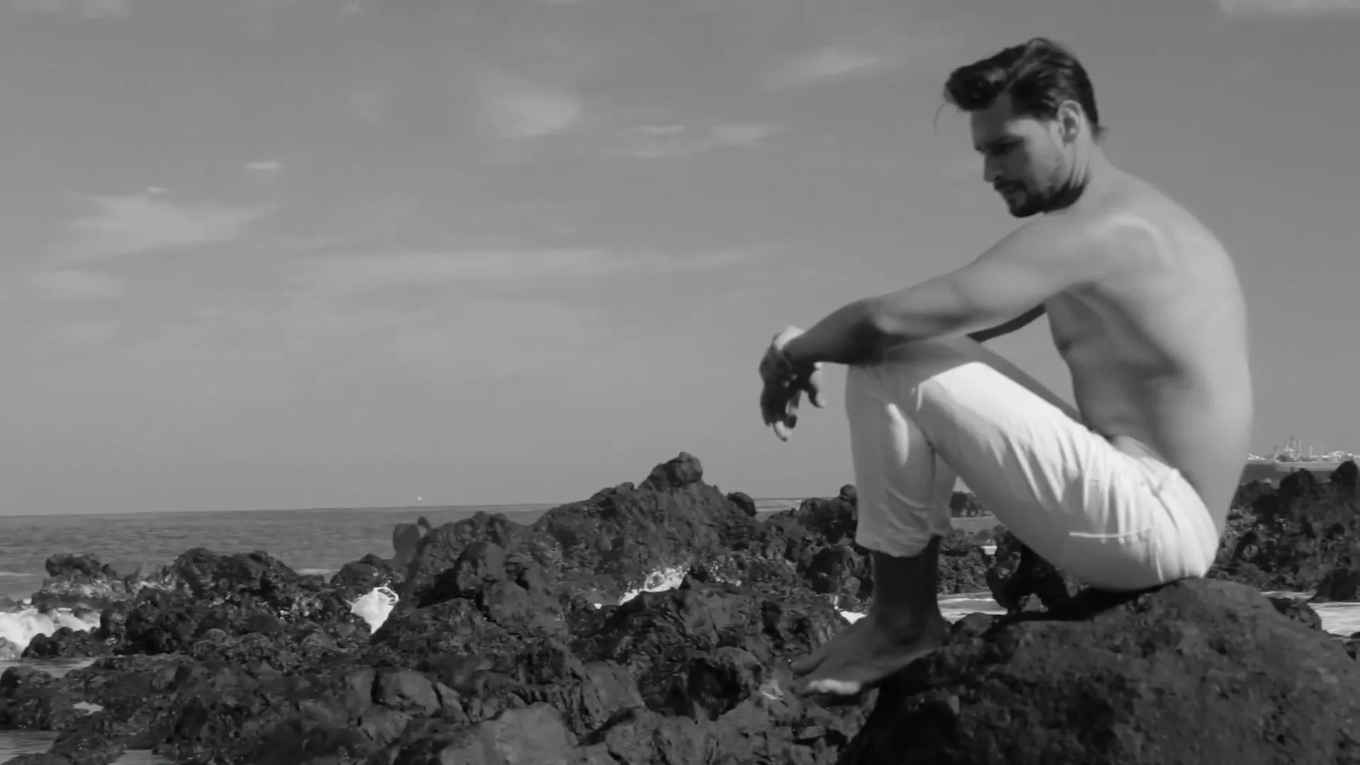 A shirtless man in white pants sits on a rocky beach, looking out at the ocean, then turns to face the camera, with a pizza superimposed over the scene.