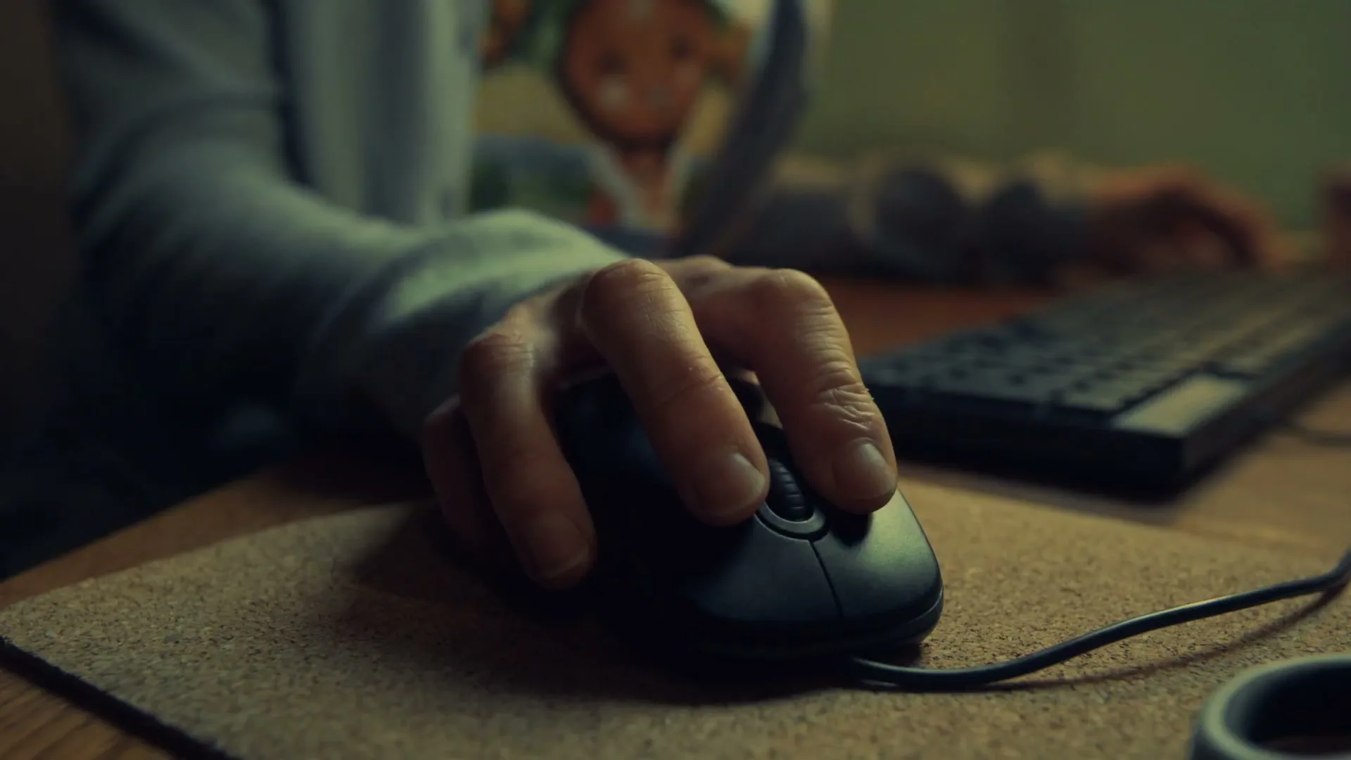 A person's hand is seen in a medium closeup, clicking a computer mouse on a cork mousepad, next to a keyboard on a wooden desk.