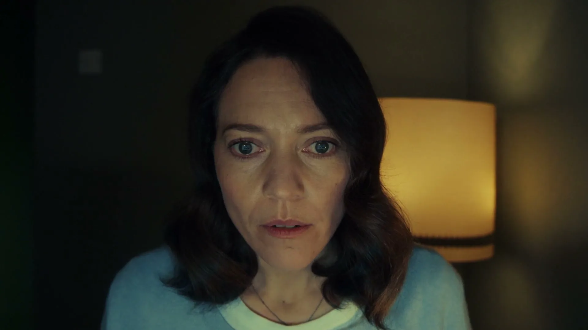 A woman with dark hair and a light blue sweater looks directly at the camera with a worried expression in a dimly lit room.
