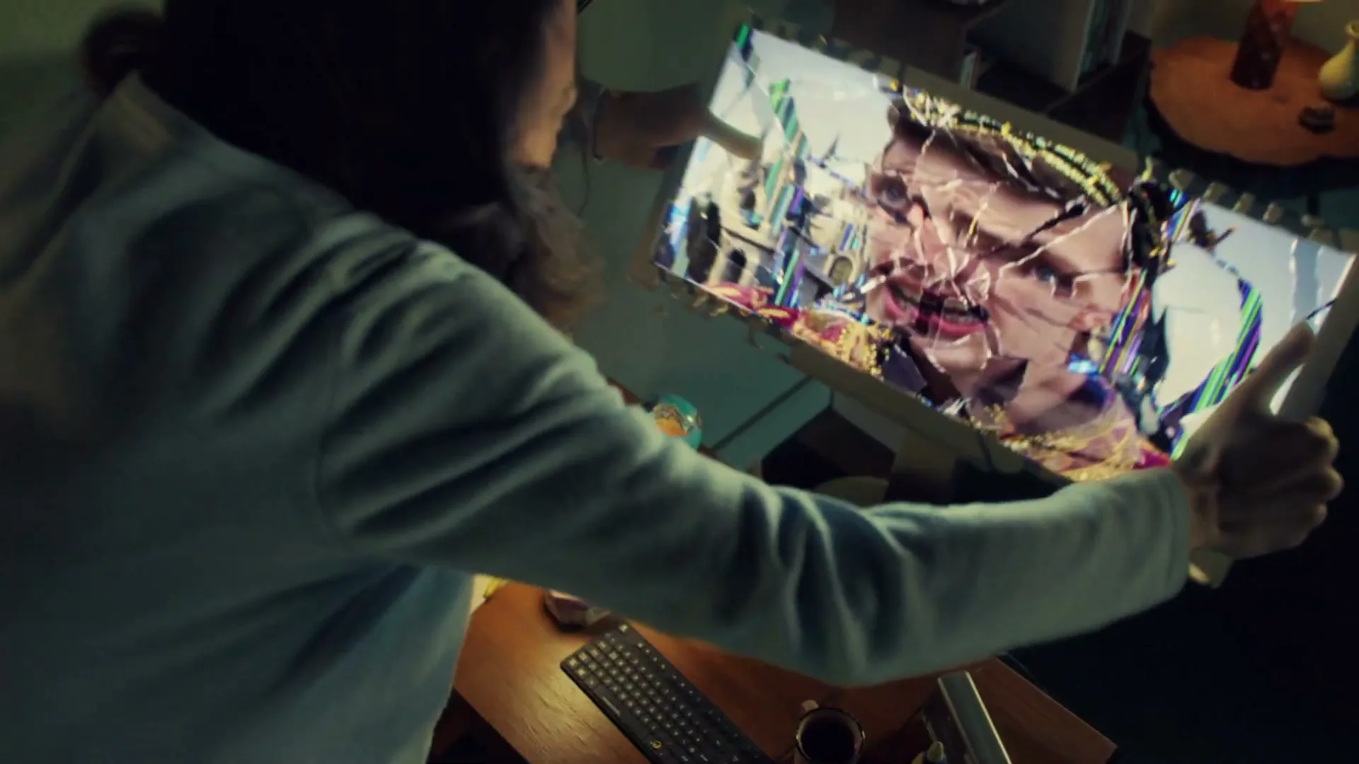 A man in a light blue shirt holds a computer monitor with both hands, as the screen shatters into colorful fragments, in a dimly lit room.