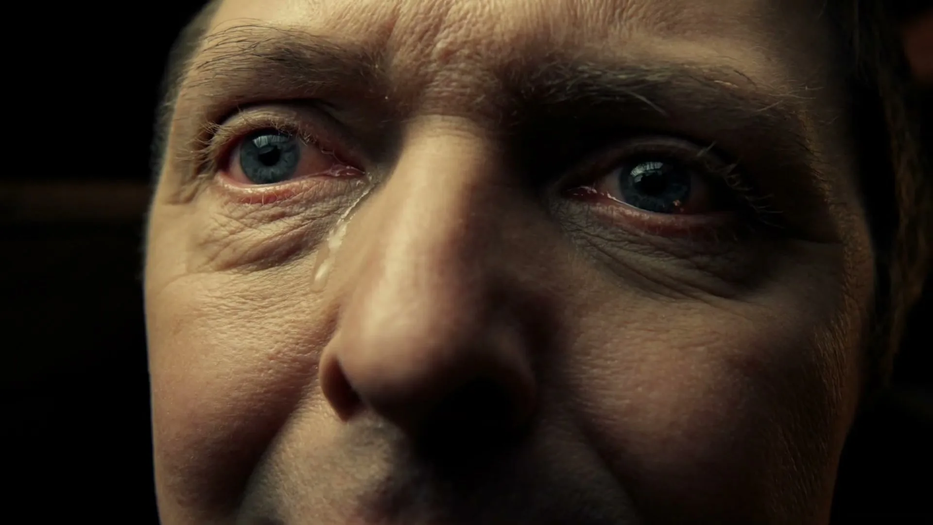 A man with blue eyes and a tear rolling down his cheek looks distressed in a dark, indeterminate interior setting.