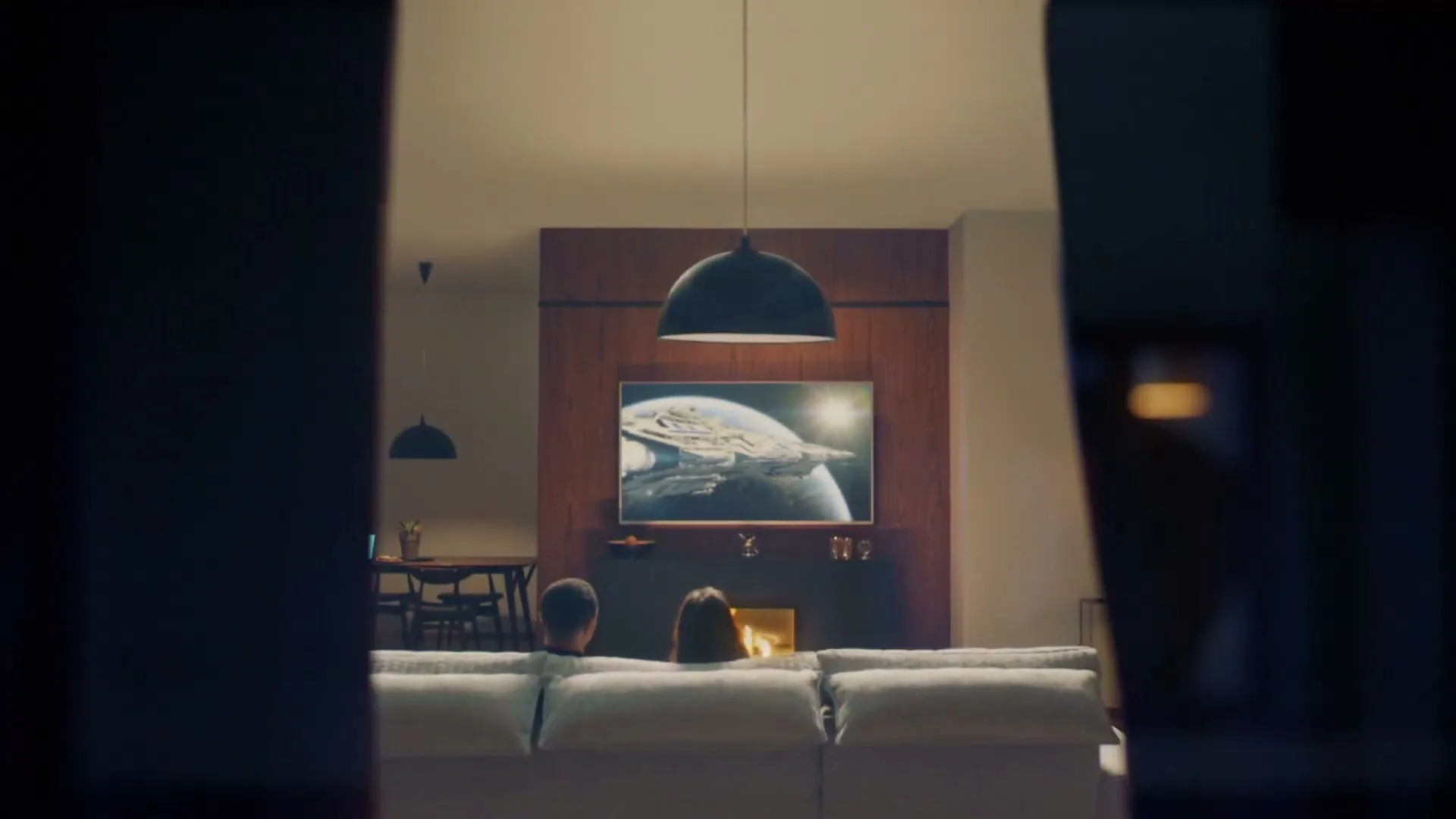 A couple sits on a sofa, watching a spaceship on a large TV screen in a modern living room, as a window slowly closes in front of them.