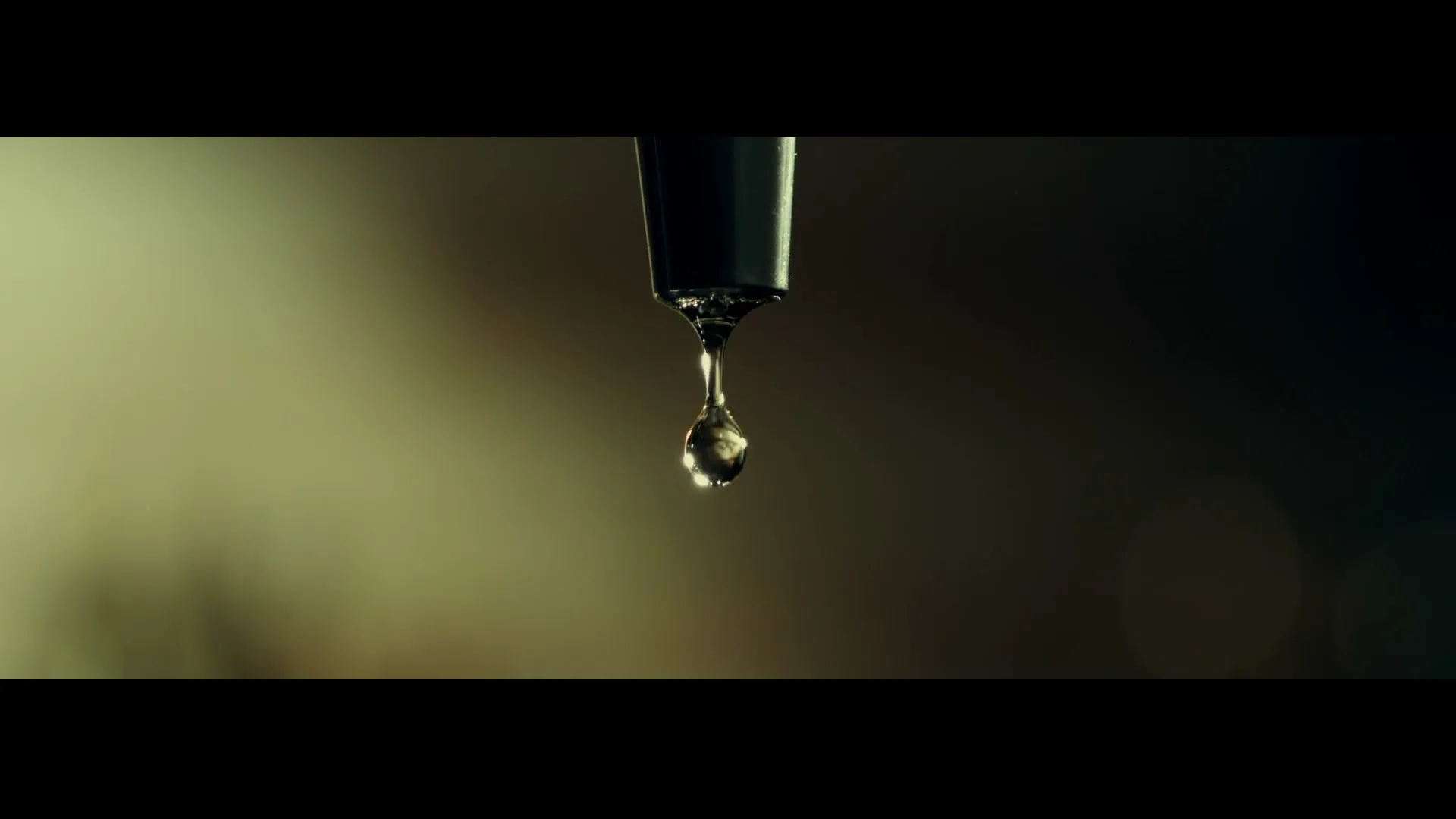 A close-up shot shows a single drop of liquid forming and dripping from a dark spout against a blurred, warm-toned background.