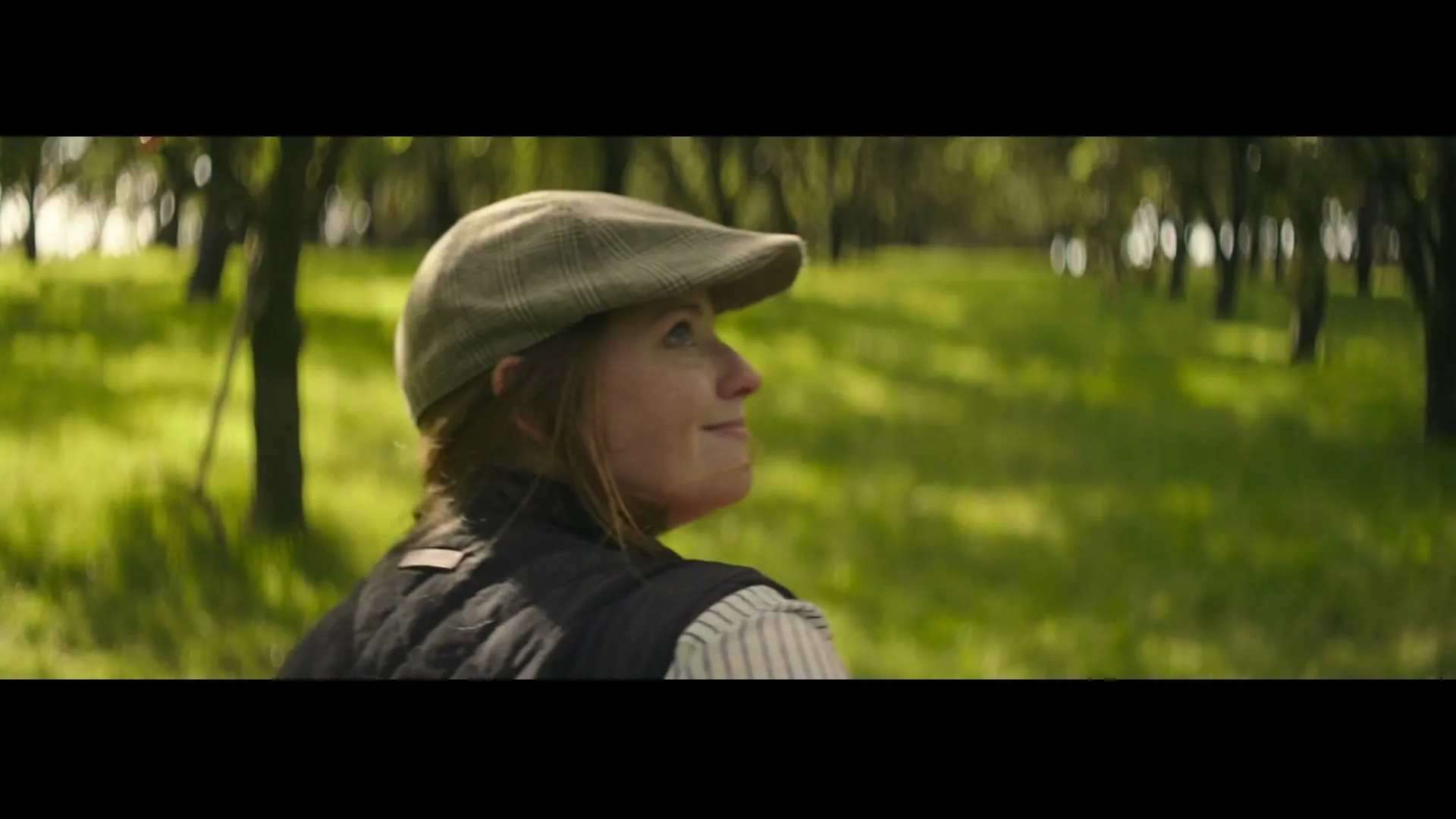 A woman in a flat cap and quilted vest smiles, looking up and to the right, in a sunny orchard.