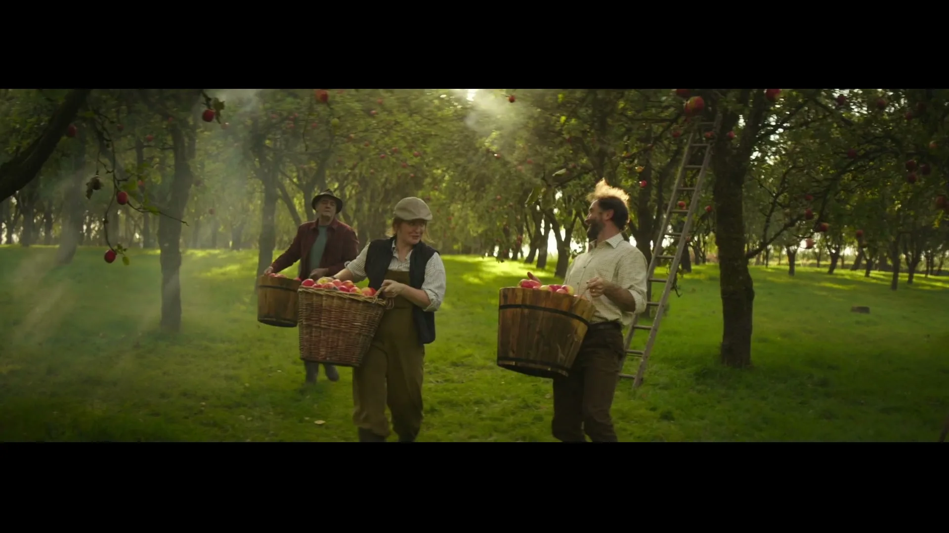 Three people, two men and one woman, are walking through an apple orchard, carrying baskets full of apples, with sunlight filtering through the trees.