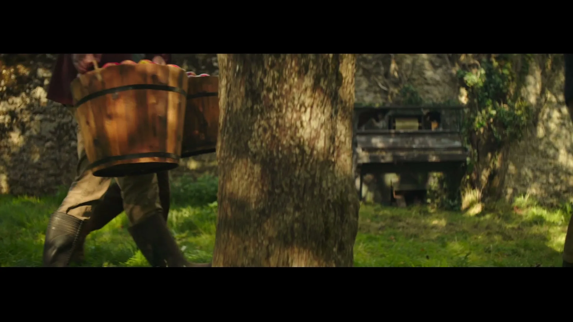 Two people in rustic clothing carry wooden buckets full of apples through a sun-dappled orchard, with an old piano visible in the background.