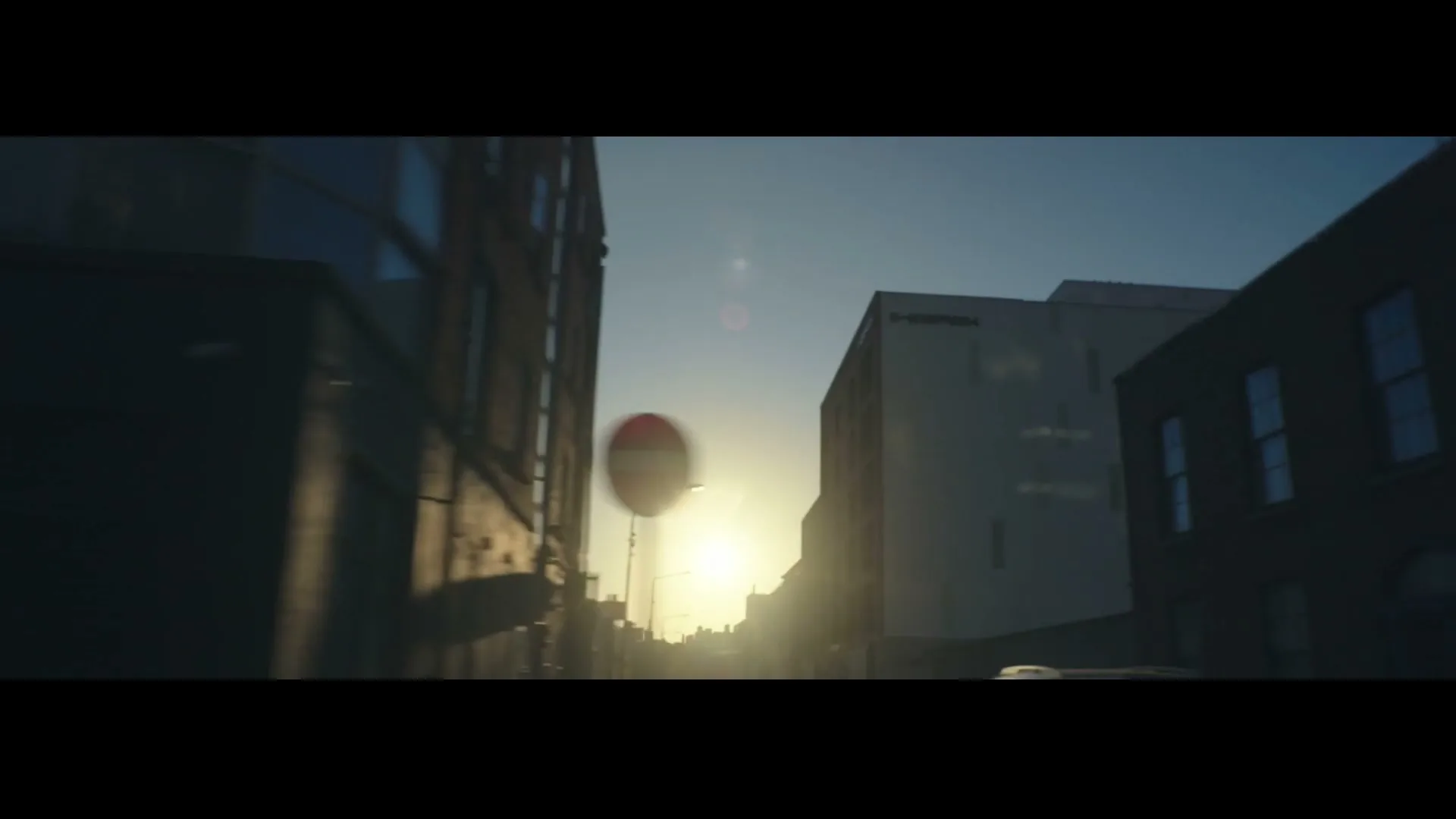 The sun sets behind buildings on a city street, with a stop sign visible on the left.