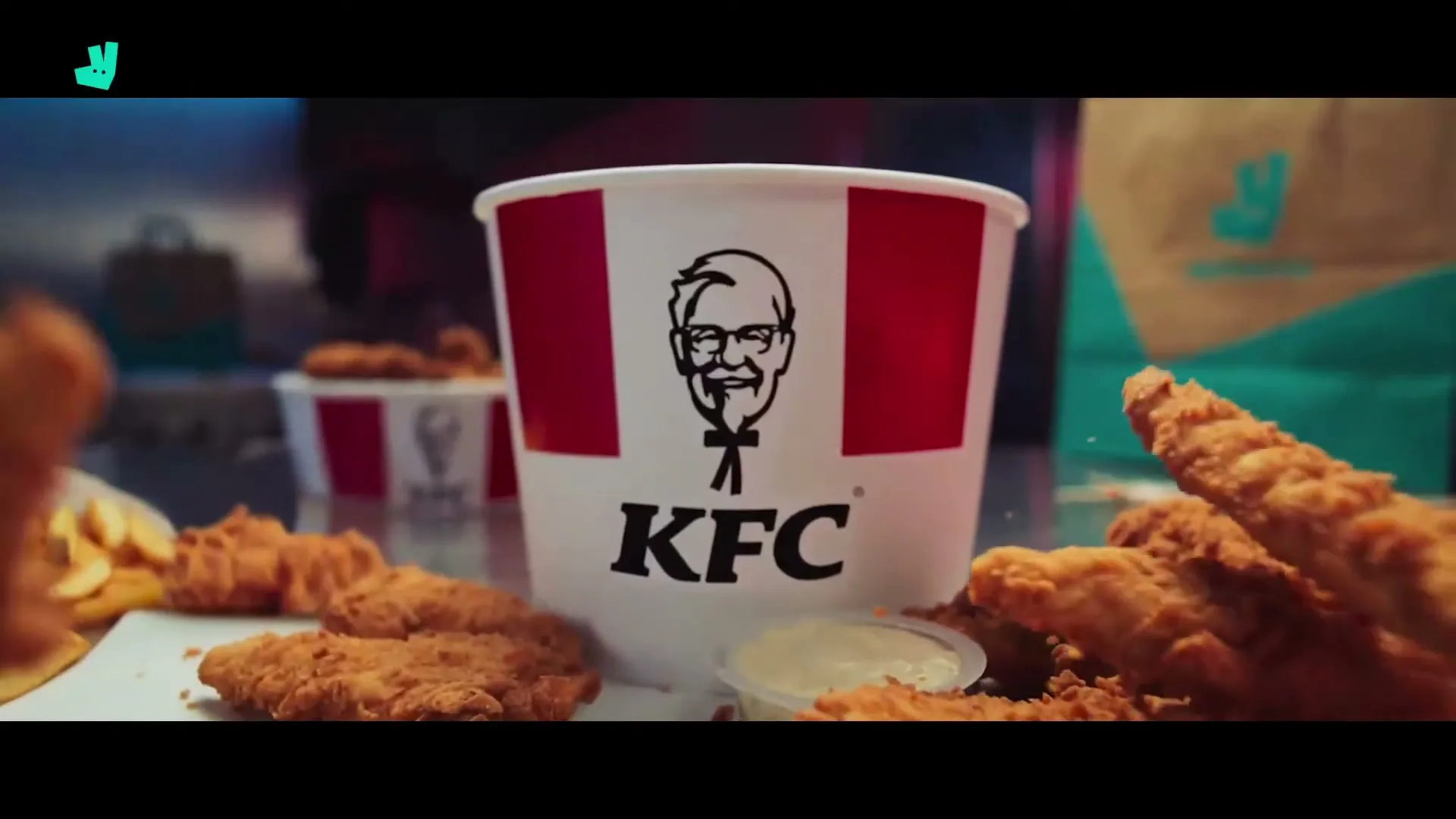 A bucket of KFC fried chicken and fries is surrounded by chicken tenders and dipping sauce on a table, with Deliveroo bags in the background.