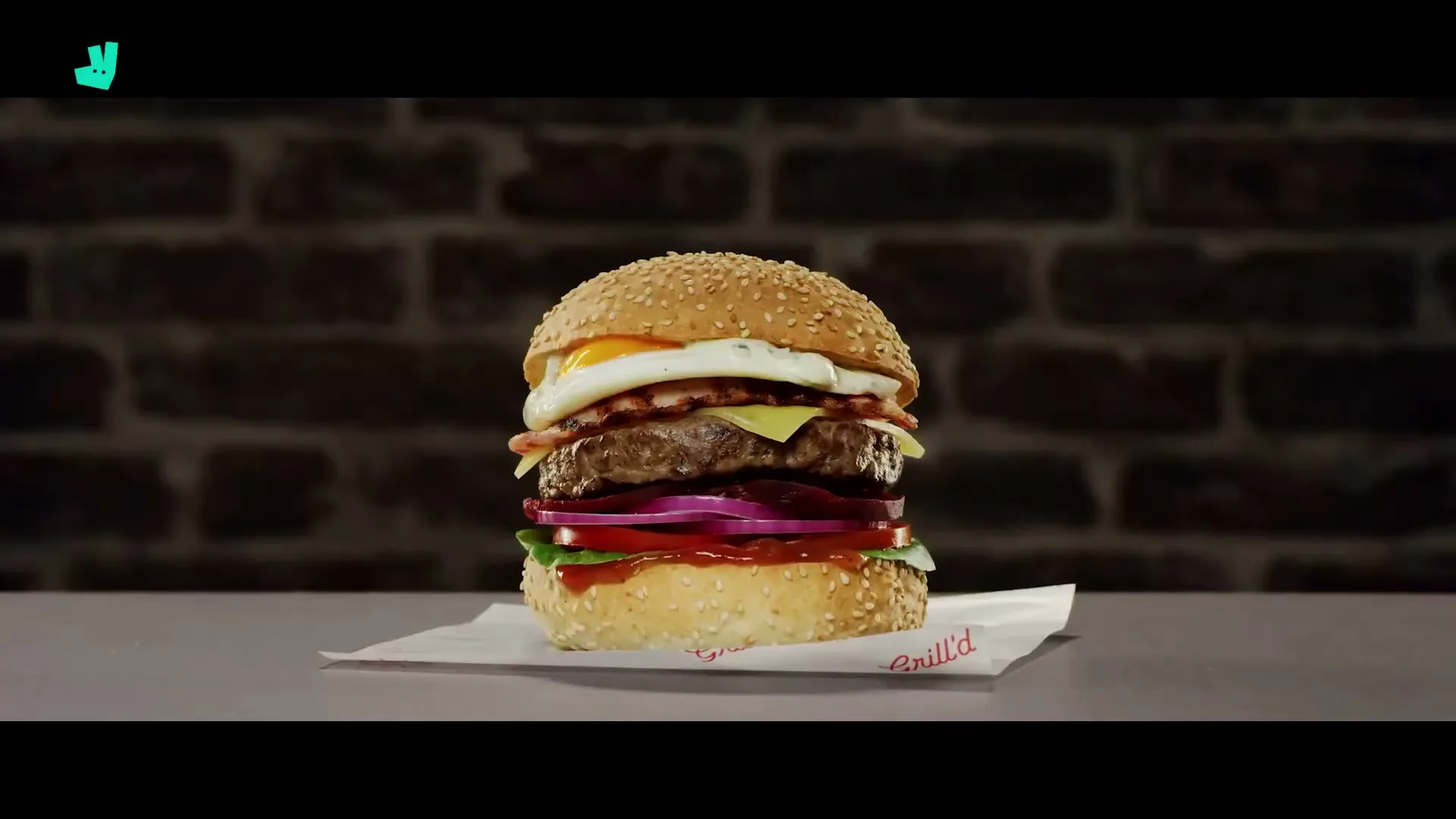 A delicious burger is assembled layer by layer on a white paper, with a brick wall background, then a Grill'd pick is added.
