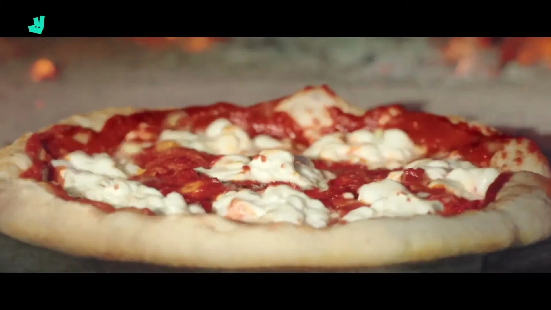 A close-up shot of a pizza with tomato sauce and mozzarella cheese cooking in a wood-fired oven, with flames visible in the background.