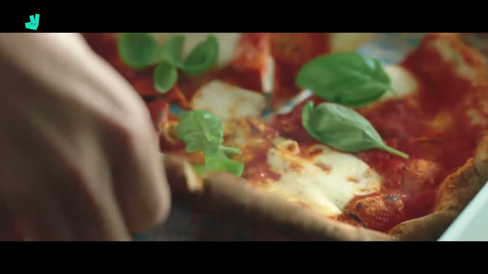 A hand reaches into a pizza box to grab a slice of freshly made pizza with basil leaves on top, in a close-up shot.