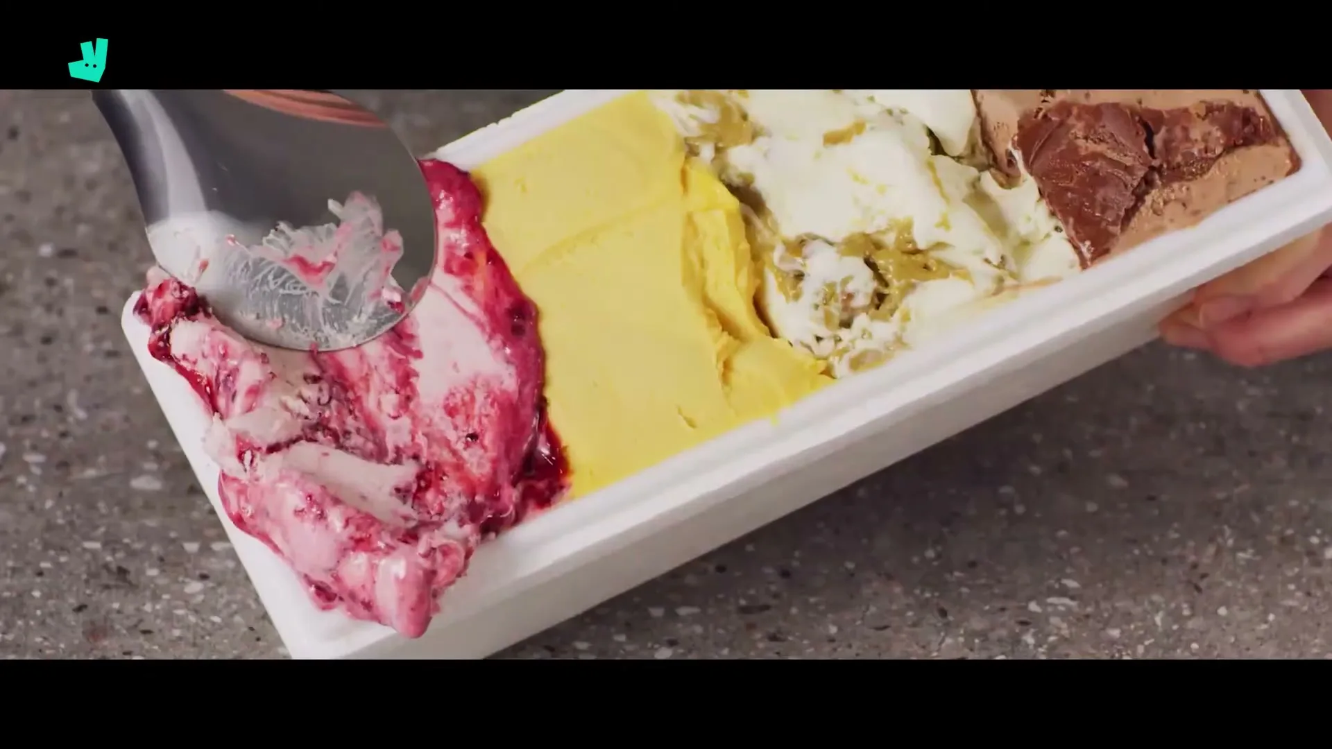 A person scoops a spoonful of raspberry ice cream from a multi-flavored tub of ice cream on a grey countertop.