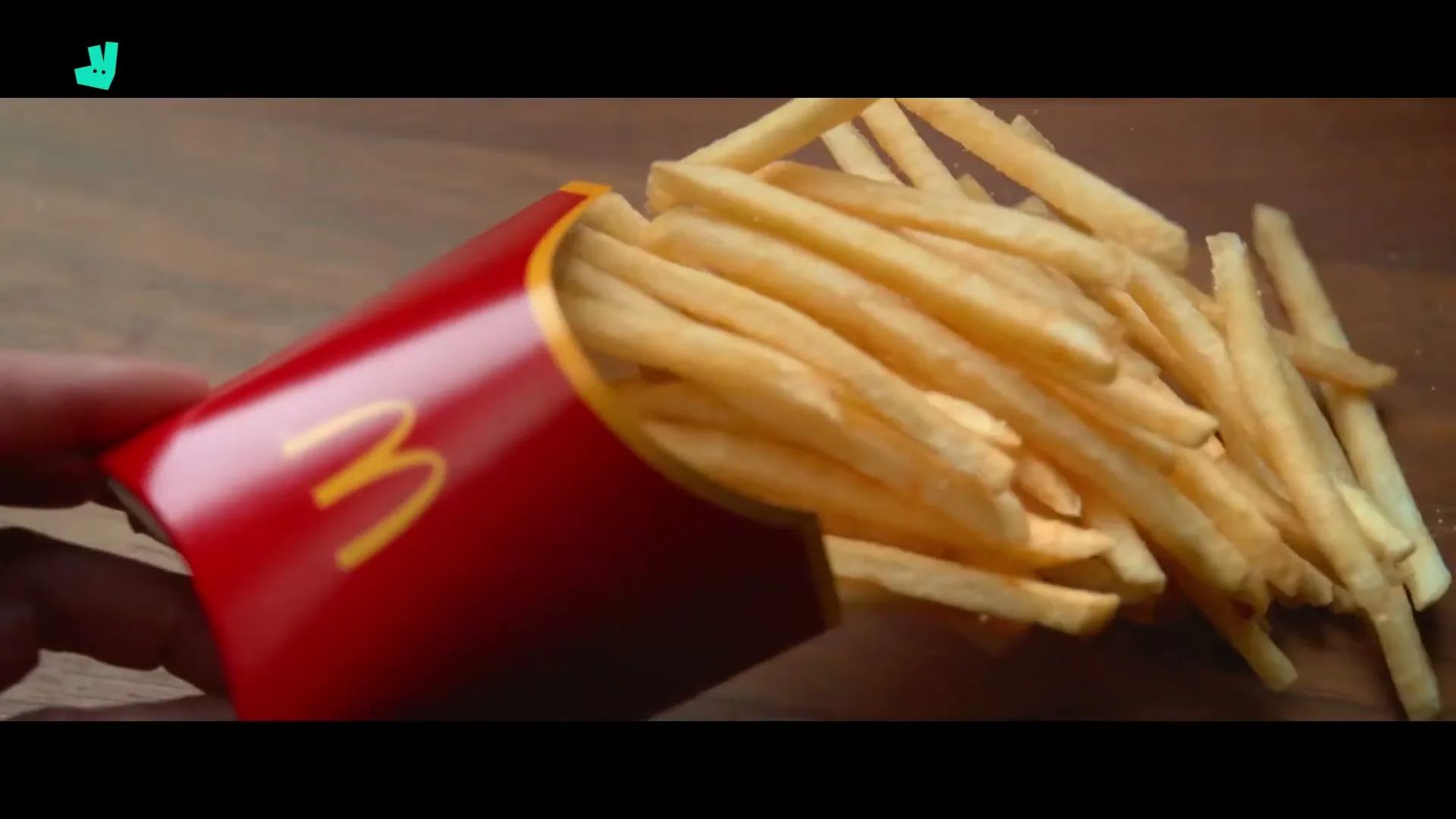 A hand holds a red McDonald's fries carton, tilting it to spill golden french fries onto a wooden surface.