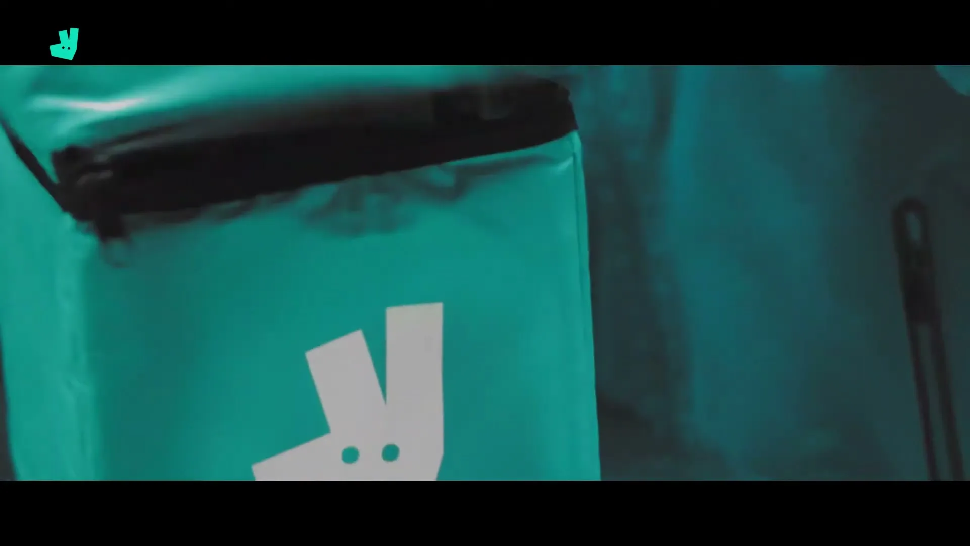 A person in a white coat places food containers into a teal Deliveroo delivery bag, which is then zipped up, revealing the brand's logo.