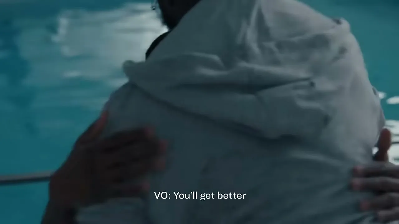 Two men embrace in a swimming pool, one comforting the other, who looks up with a pained expression.