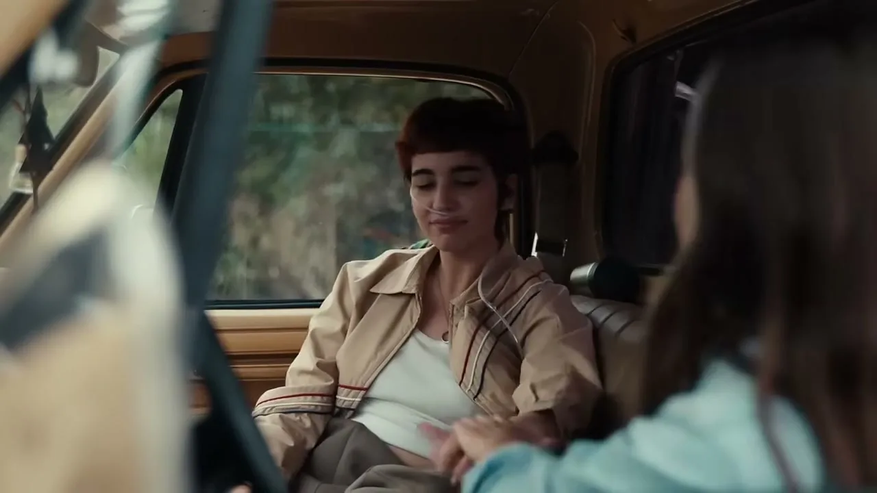 A young woman with short hair and a nasal cannula smiles and holds hands with another woman in the passenger seat of a vintage truck.