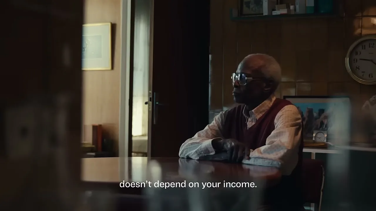 An older Black man with glasses and a maroon vest sits at a table, looking off-screen in a dimly lit room.
