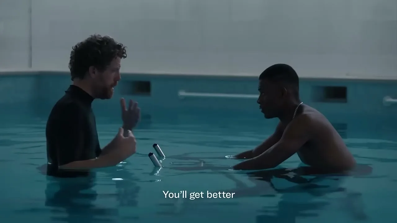 A male physical therapist with curly hair and a black shirt talks to a young male patient in a swimming pool, who is using an underwater exercise bike.