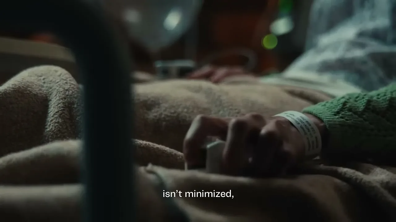 A patient's hand with an ID bracelet and pulse oximeter lies on a blanket in a hospital bed, with medical equipment visible in the background.