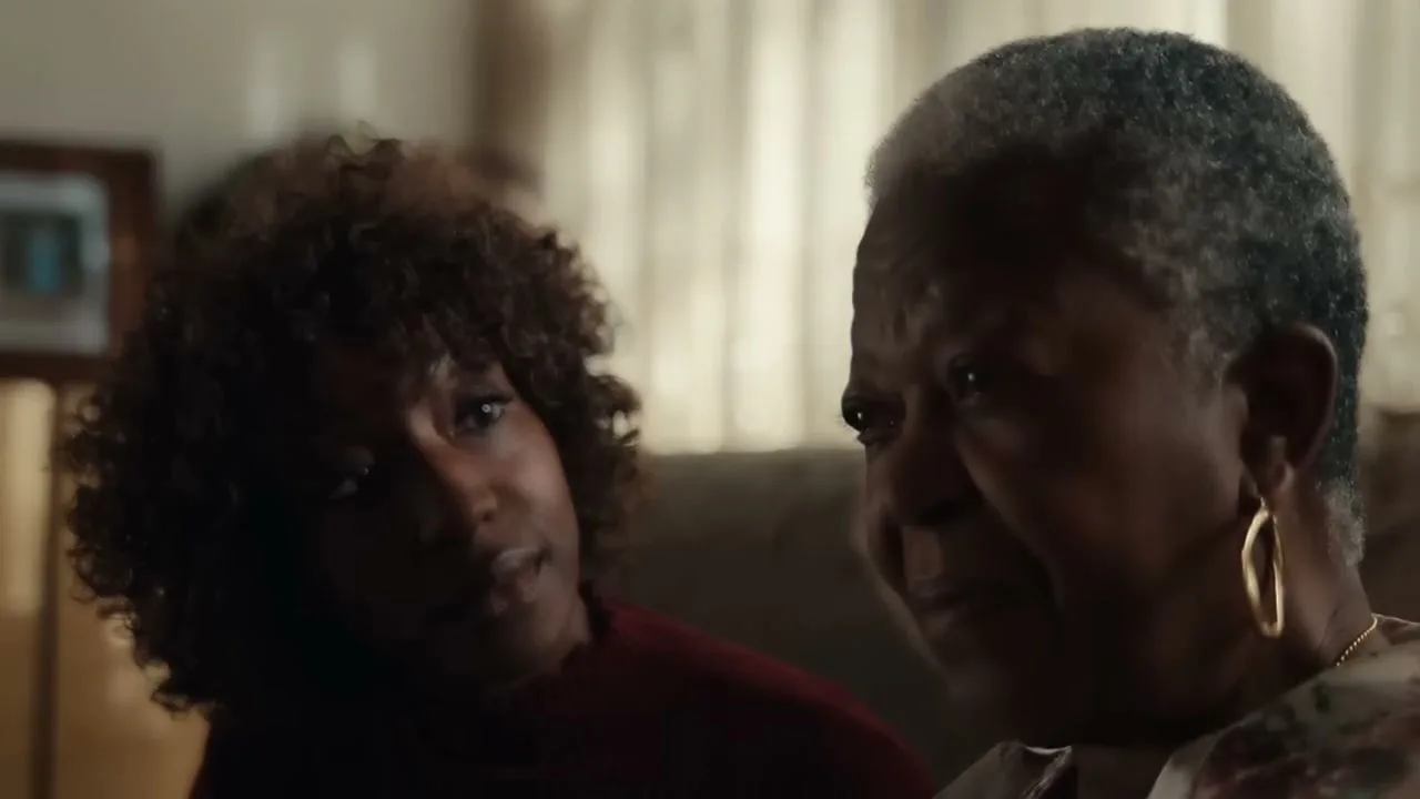 A younger Black woman with curly hair looks at an older Black woman with gray hair, who looks away, in a living room.