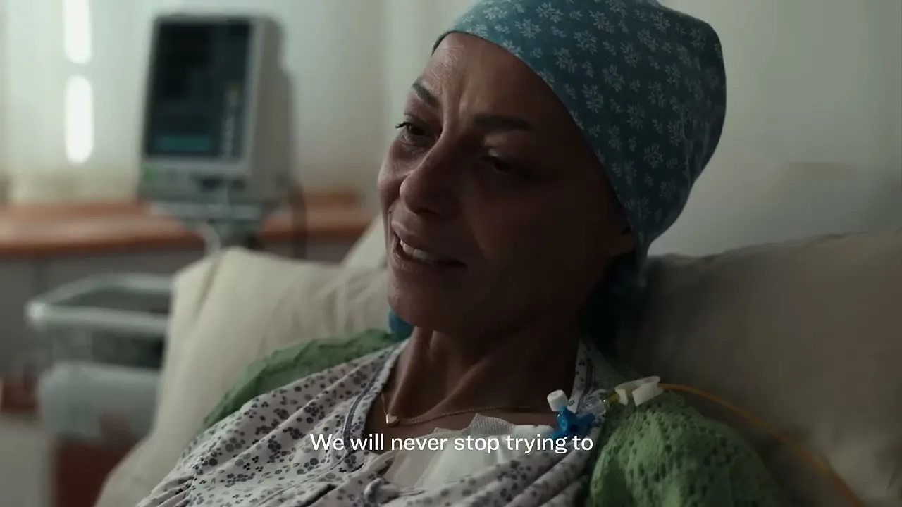 A female patient with a headscarf and an IV drip looks distressed while lying in a hospital bed.