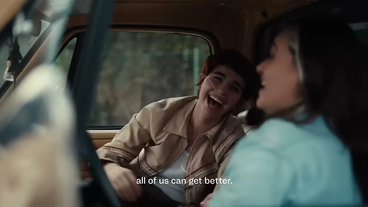 A young person with a nasal cannula laughs joyfully while sitting in the passenger seat of a car, talking to another person.