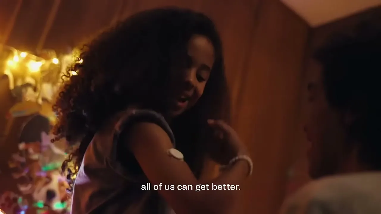 A young girl with curly hair, wearing a medical device on her arm, smiles while looking at someone off-screen in a warmly lit room.