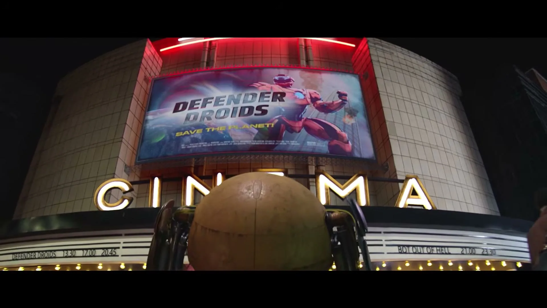 A large, round, brown robot with green legs stands in front of a cinema at night, with a movie poster for "Defender Droids" visible above.