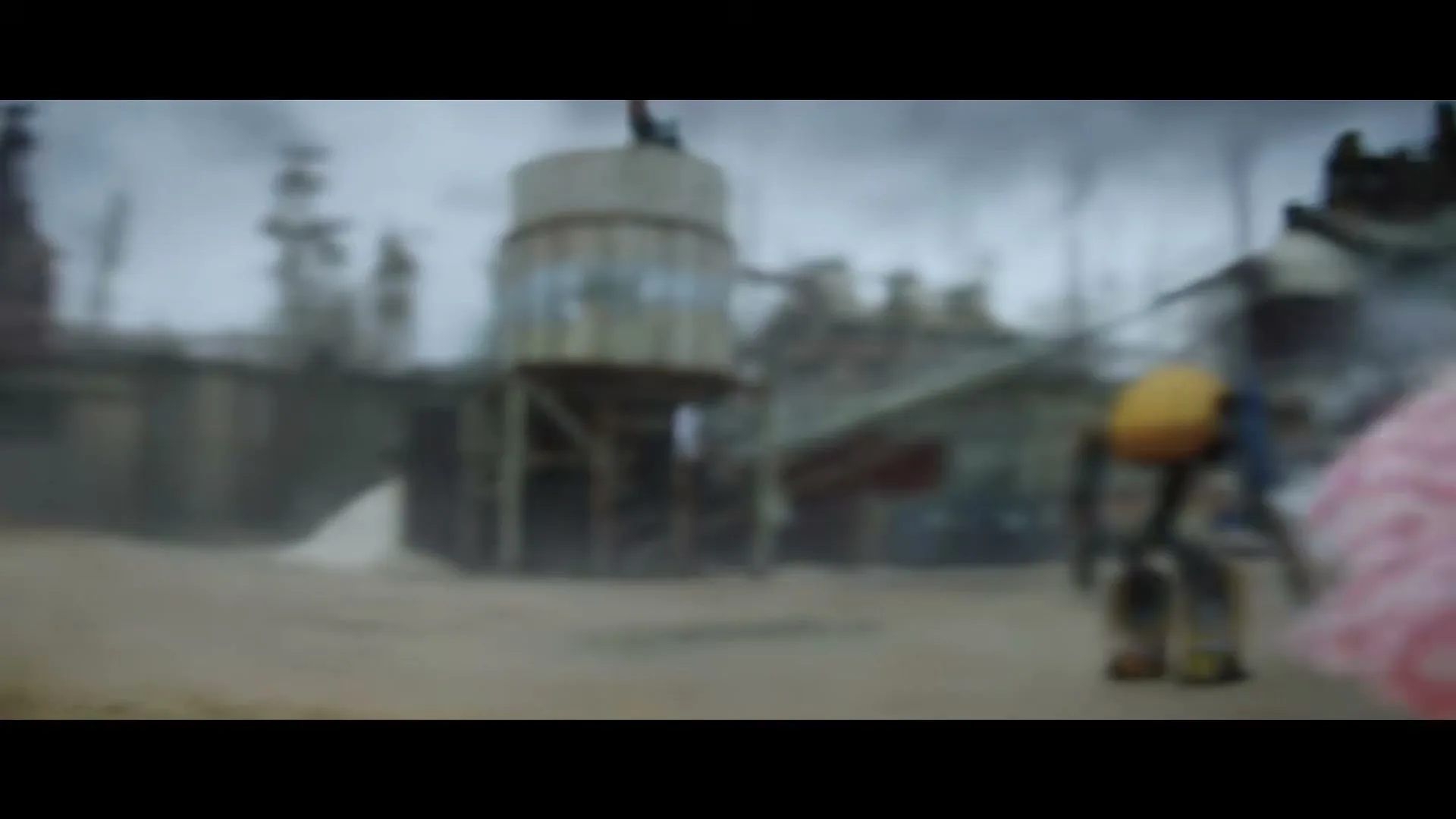 A small, yellow and grey robot walks away from the camera towards a large, industrial factory under a cloudy sky.