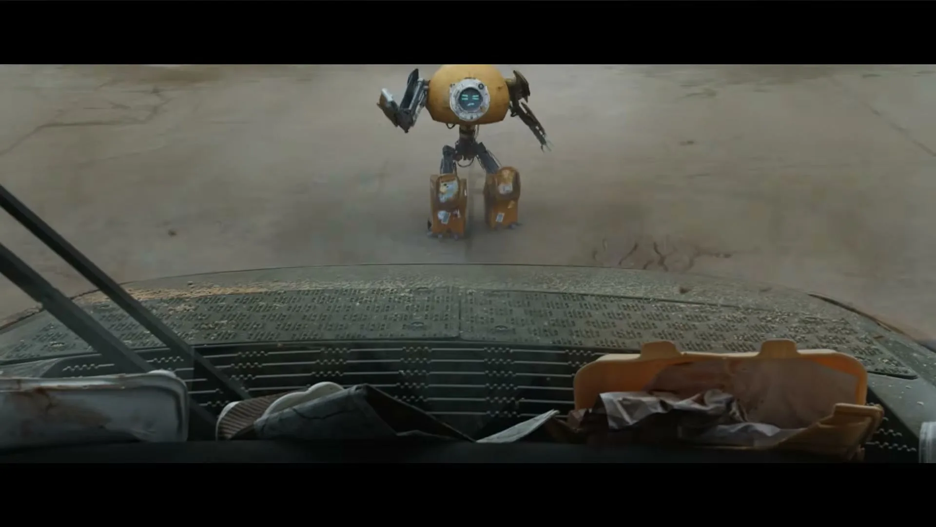 A small, yellow, round robot with thin limbs stands on a cracked concrete surface, viewed from behind a car's dashboard.
