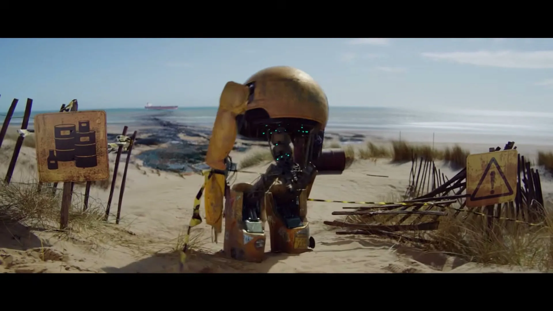 A small, rusty, yellow robot with a domed head and glowing blue lights walks on a sandy beach with warning signs and a ship in the distance.