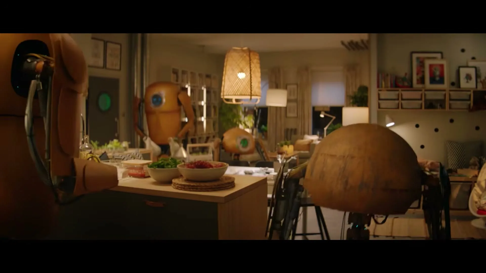 A group of brown, spherical robots with glowing eyes are gathered around a kitchen island, preparing food in a cozy, well-lit home.