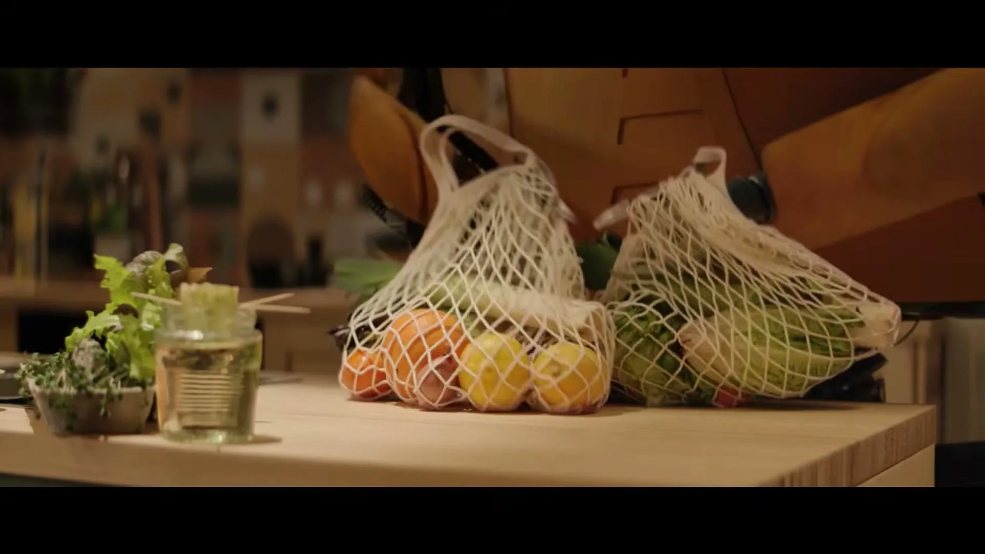A robot arm places two mesh bags of fresh produce onto a wooden kitchen counter, next to a small plant in a jar.
