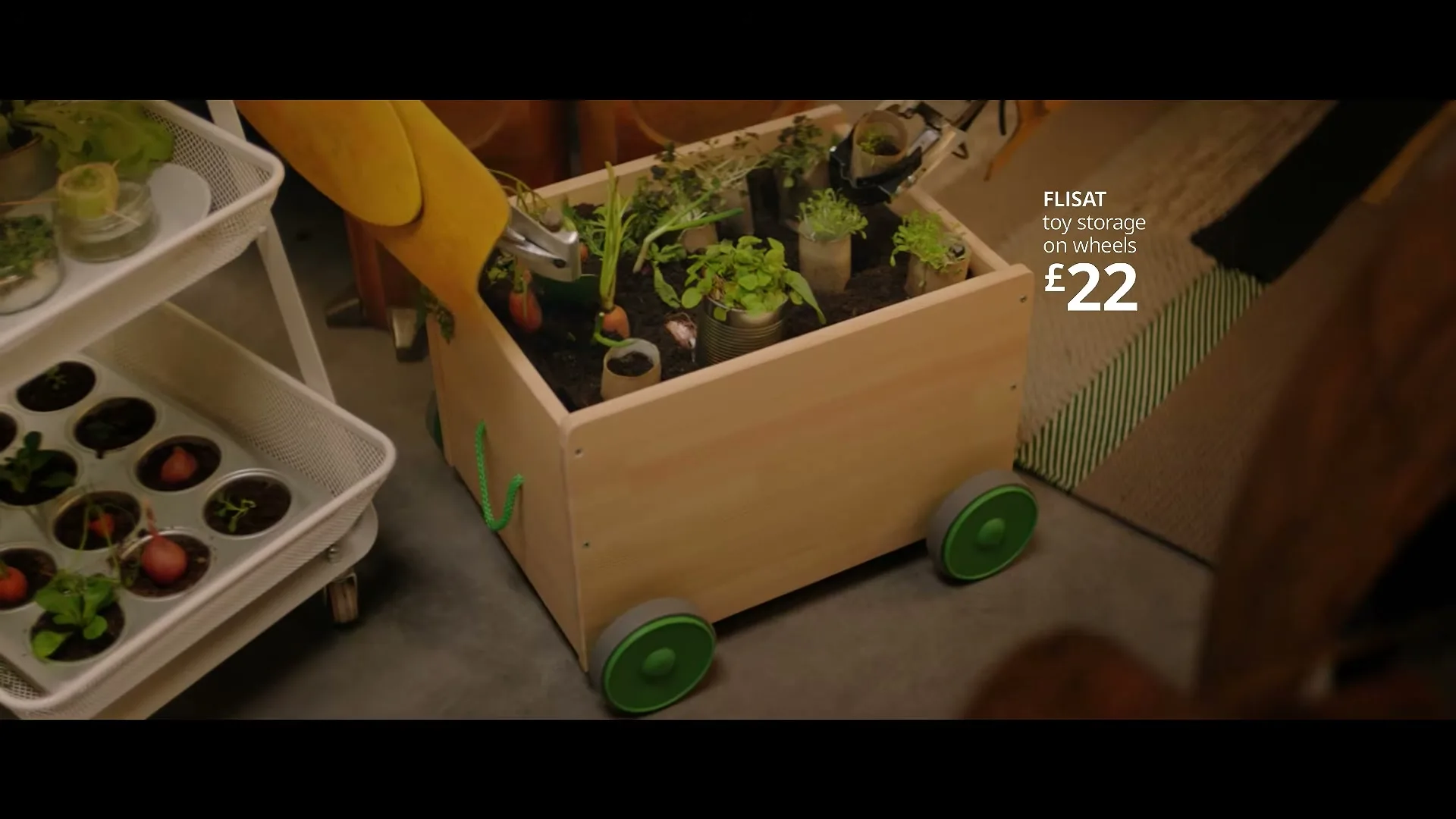 A wooden toy storage cart on wheels, repurposed as a planter with various small plants, sits on a concrete floor next to a white metal shelving unit with more plants.