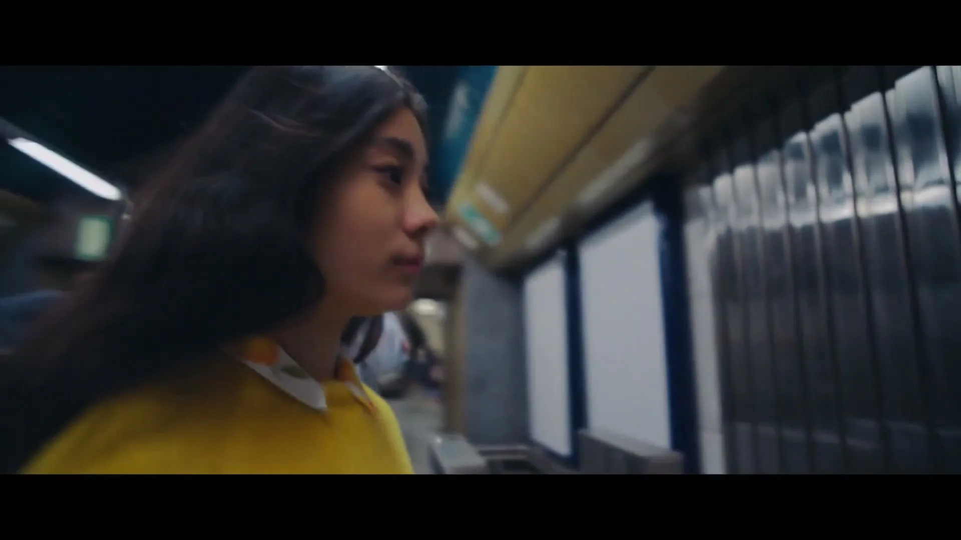 A young woman with long dark hair, wearing a yellow sweater, walks through a busy subway station, passing through turnstiles and looking around.