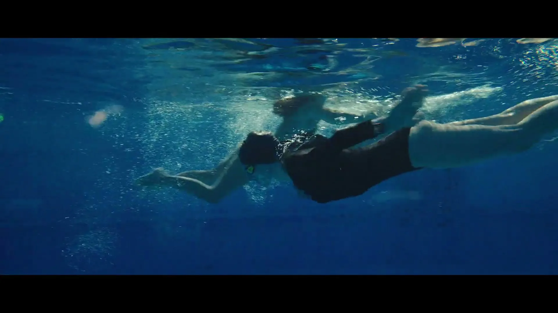 A woman in a dark swimsuit and goggles swims underwater in a pool, with other swimmers and the pool edge visible in the background.