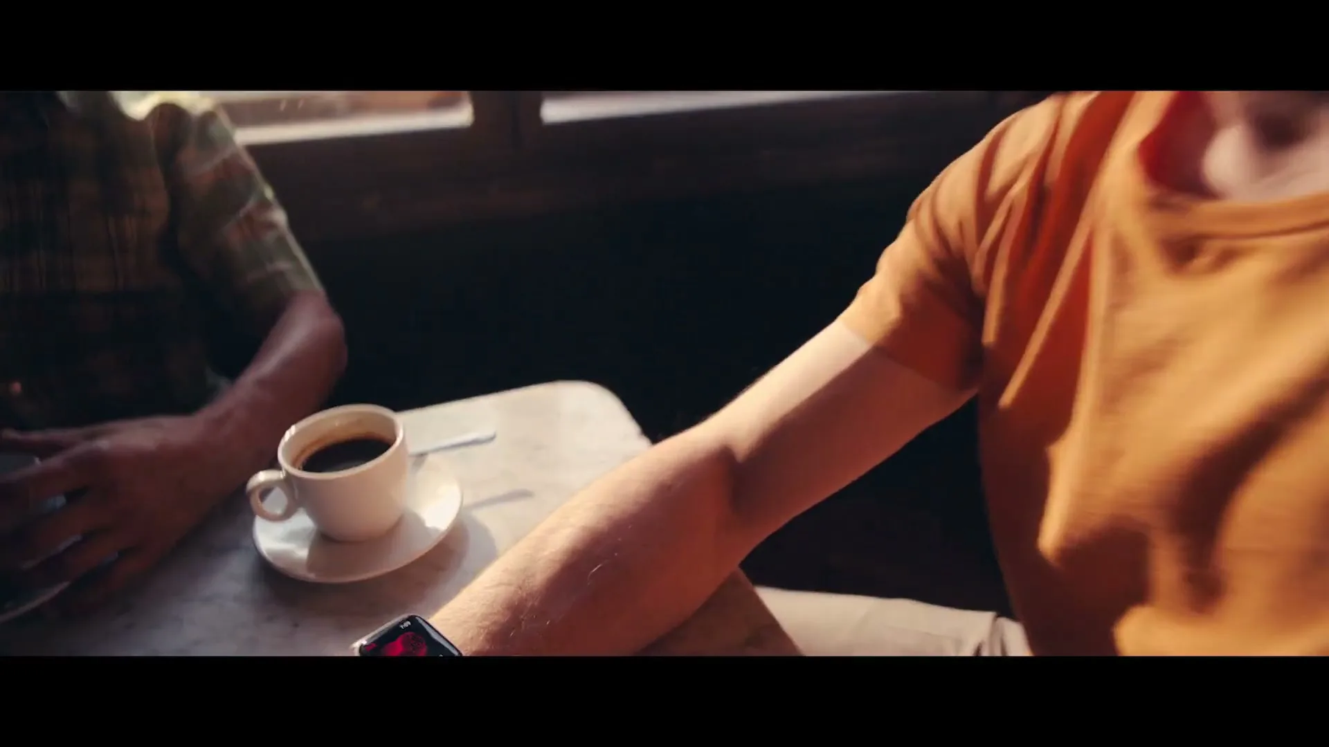 A man in an orange shirt sits at a cafe table, checking his Apple Watch, which displays a heart rate monitor, then transitions to a CG animation of a human heart.