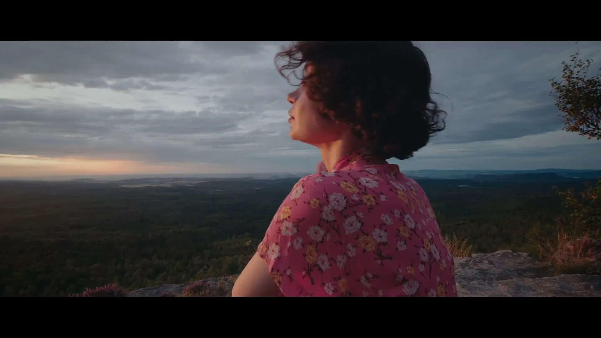 A young woman with dark curly hair, wearing a pink floral dress, sits on a rocky cliff edge, looking out at a vast forest landscape at sunset.