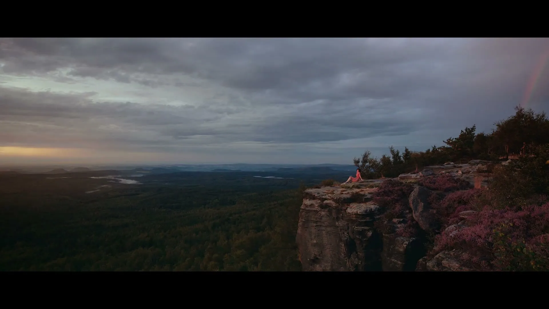 A woman in a pink top sits on a cliff edge, looking out over a vast forest landscape under a cloudy sky with a faint rainbow.