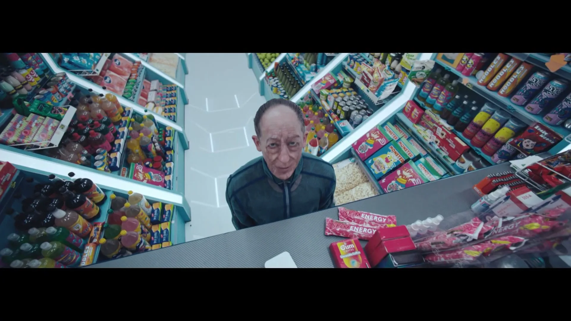 A man with a wrinkled face and receding hairline looks up at the camera from behind a counter in a brightly lit, futuristic convenience store.