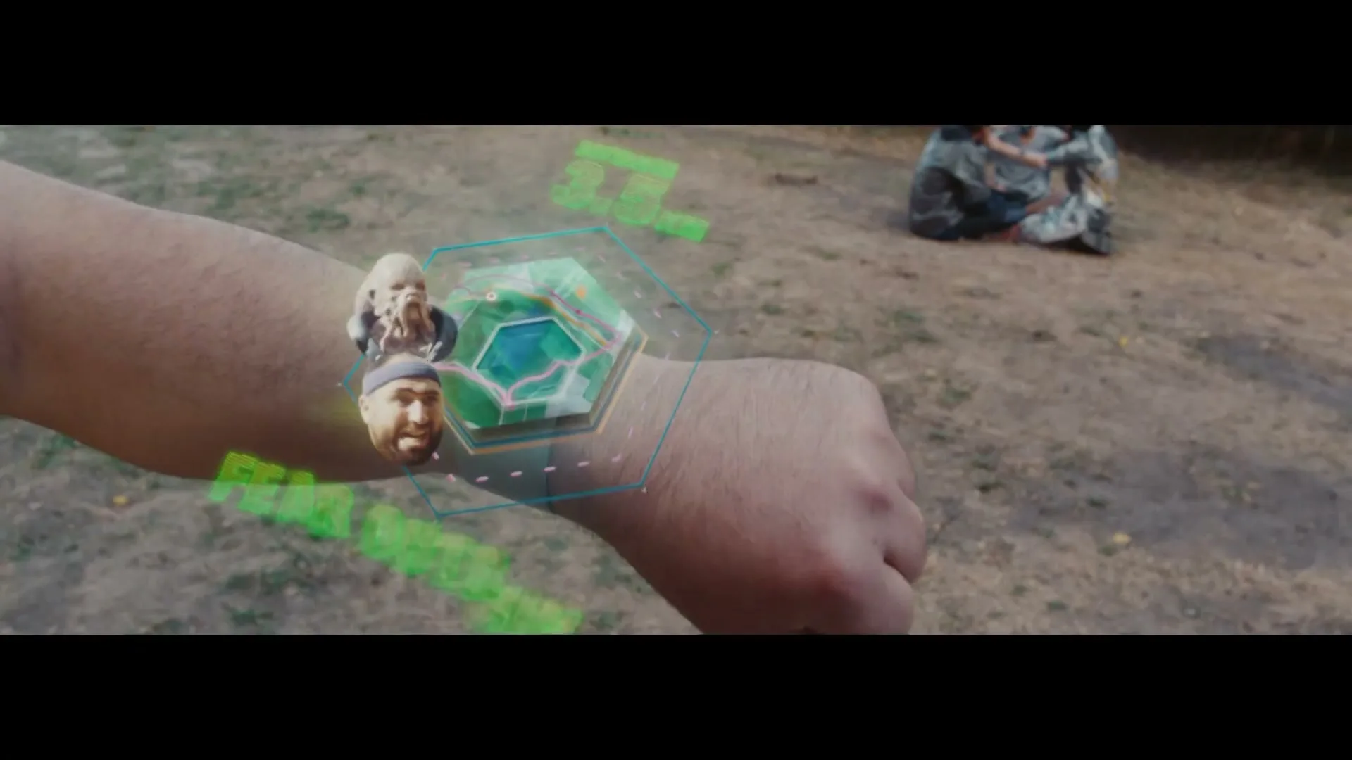 A person's arm with a holographic display showing a map and two faces, with a blurred background of dry grass and a camouflage-covered object.