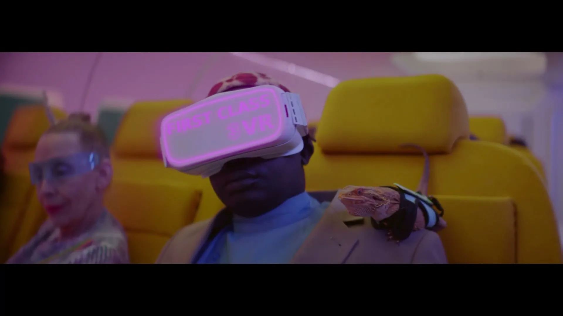 A man wearing a VR headset with glowing text sits on an airplane next to an older woman, with a lizard on his shoulder.