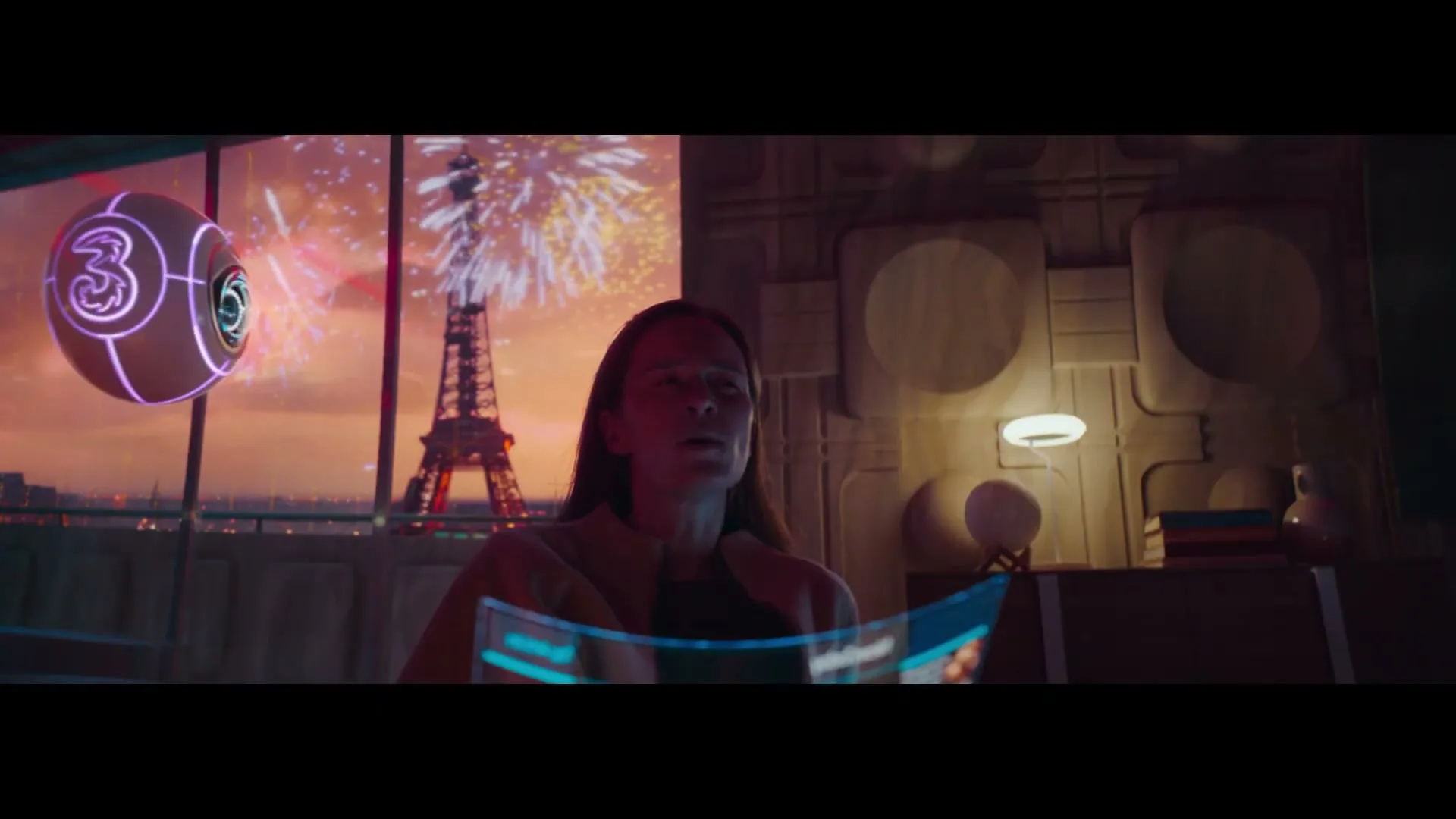 A woman in her 30s looks at a holographic screen while a glowing orb floats beside her, with the Eiffel Tower and fireworks visible outside a window.