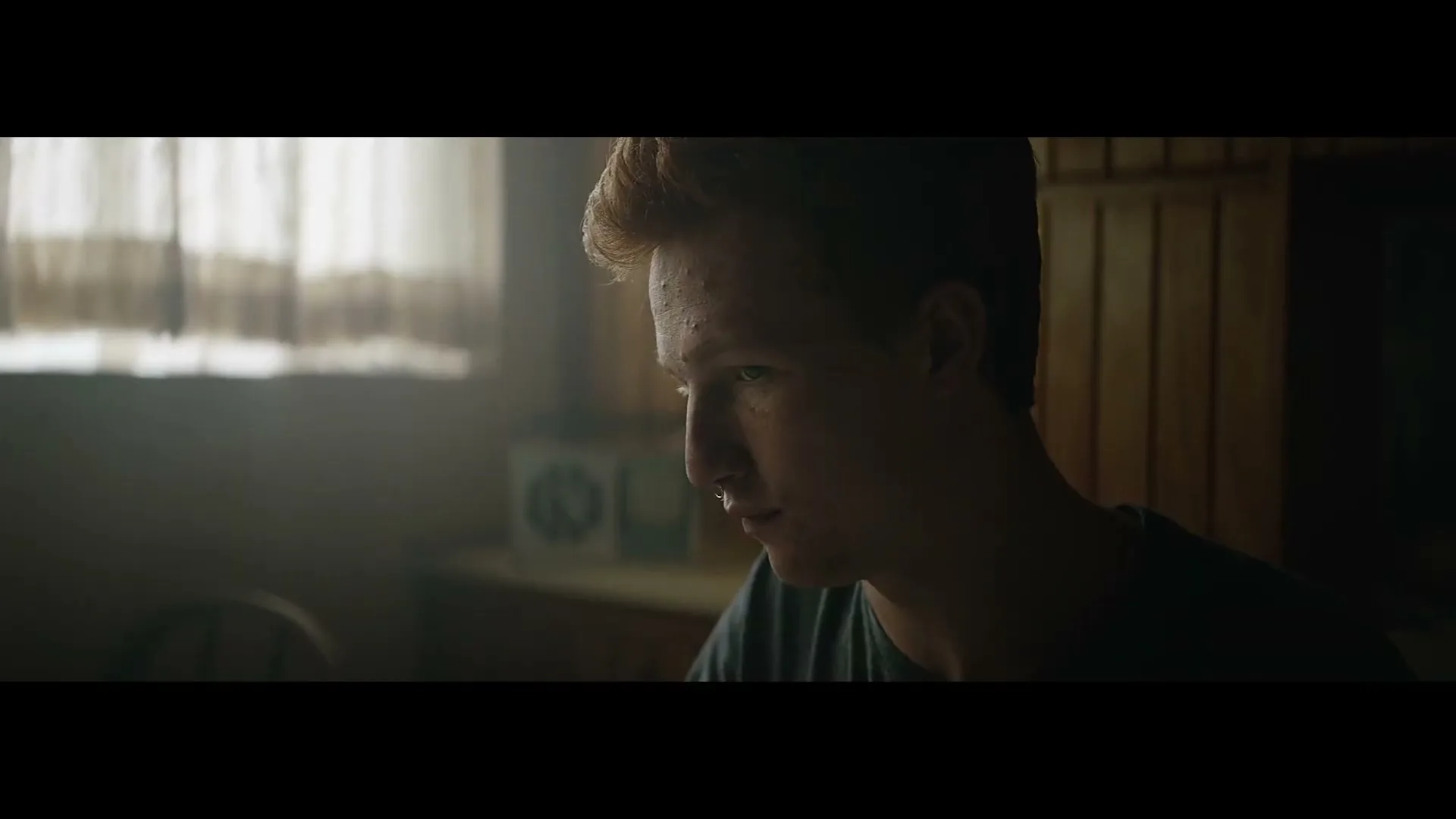 A young man with red hair and a nose ring looks down thoughtfully, illuminated by a window in a dimly lit room.